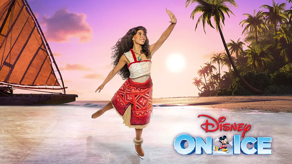 An ice skater dressed as Moana glides along the ice with her hand in the air. The background is a beach setting complete with a shoreline, palm trees, and a sunset turning the sky a purplish-orange hue. In the far background is Moana’s immediately recognizable boat.