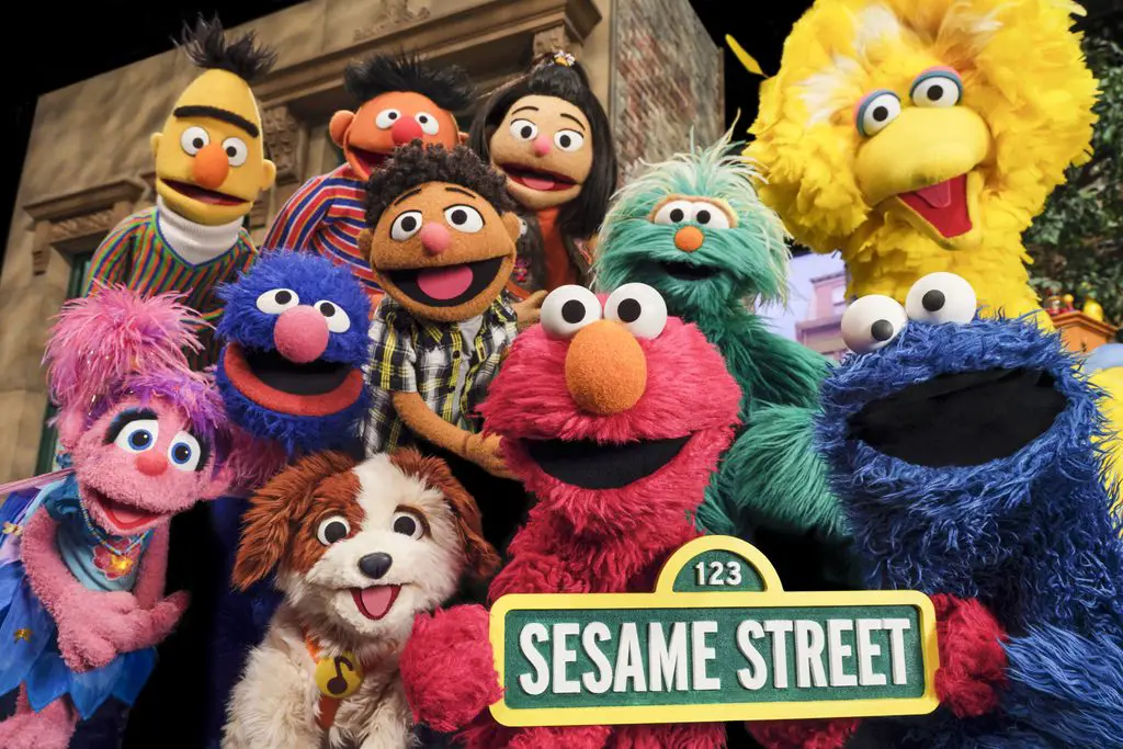 "Group of colorful Sesame Street puppet characters outdoors smiling together, holding a Sesame Street sign, with brownstone buildings and greenery in the background."